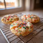 Three freshly baked cake batter cookies loaded with rainbow sprinkles cooling on a wire rack near a window.