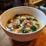 Close-up of a steaming white bowl filled with Tuscan white bean soup, featuring cannellini beans, kale, and sausage.