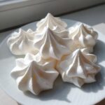 A close-up of five perfectly piped, star-shaped white meringue cookies resting on a white plate near a window.