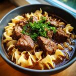 Close-up of a dark bowl filled with rich beef noodle soup, featuring tender beef chunks and egg noodles, topped with fresh parsley.