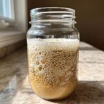 A highly active sourdough starter showing significant bubbling and foam development inside a clear glass jar.