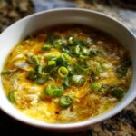 A close-up of a white bowl filled with steaming egg drop soup, topped generously with bright green sliced scallions.