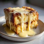 Close-up of two stacked squares of golden brown bread pudding generously covered in creamy vanilla sauce.