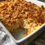 A close-up of a baked hot chicken salad casserole in a white dish with a serving removed, showing the creamy interior and crunchy topping.