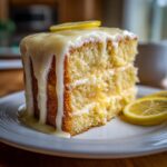 A moist slice of two-layer lemon cake recipe covered in thick white glaze and garnished with fresh lemon slices.