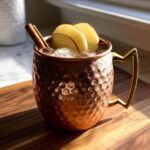A refreshing apple cider moscow mule served over ice in a hammered copper mug, garnished with apple slices and a cinnamon stick.