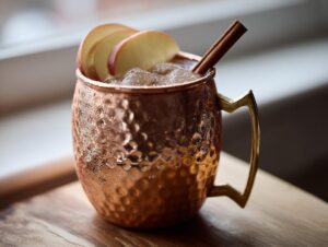 Close-up of a frosty copper mug filled with an apple cider moscow mule, garnished with apple slices and a cinnamon stick.