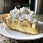 A generous slice of banana cream pie recipe featuring a graham cracker crust, creamy yellow filling, fresh banana slices, and a large dollop of whipped cream.