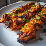 Close-up of perfectly grilled bang bang chicken skewers drizzled with orange sauce and topped with sesame seeds and green onions.
