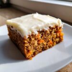 A single, moist square of carrot cake bars topped with thick cream cheese frosting on a white plate.