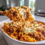 A wooden spoon lifts a cheesy, saucy portion of enchilada pasta out of a white baking dish.