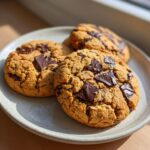 Three freshly baked, thick pudding cookies loaded with dark chocolate chunks resting on a light ceramic plate.