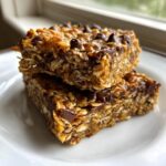 Two chewy homemade granola bars stacked, featuring oats and melted chocolate chips, resting on a white plate.
