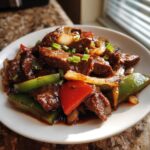 Close-up of glossy Chinese pepper steak with beef, green peppers, red peppers, and onions, garnished with scallions.