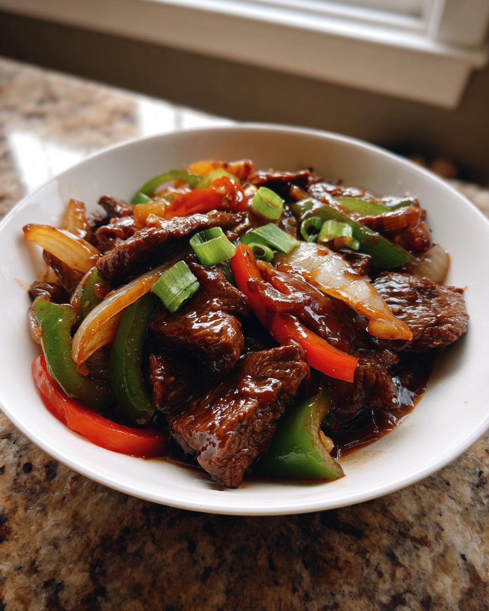 Close-up of a white bowl filled with glistening Chinese pepper steak, mixed with green bell peppers, red peppers, and onions.