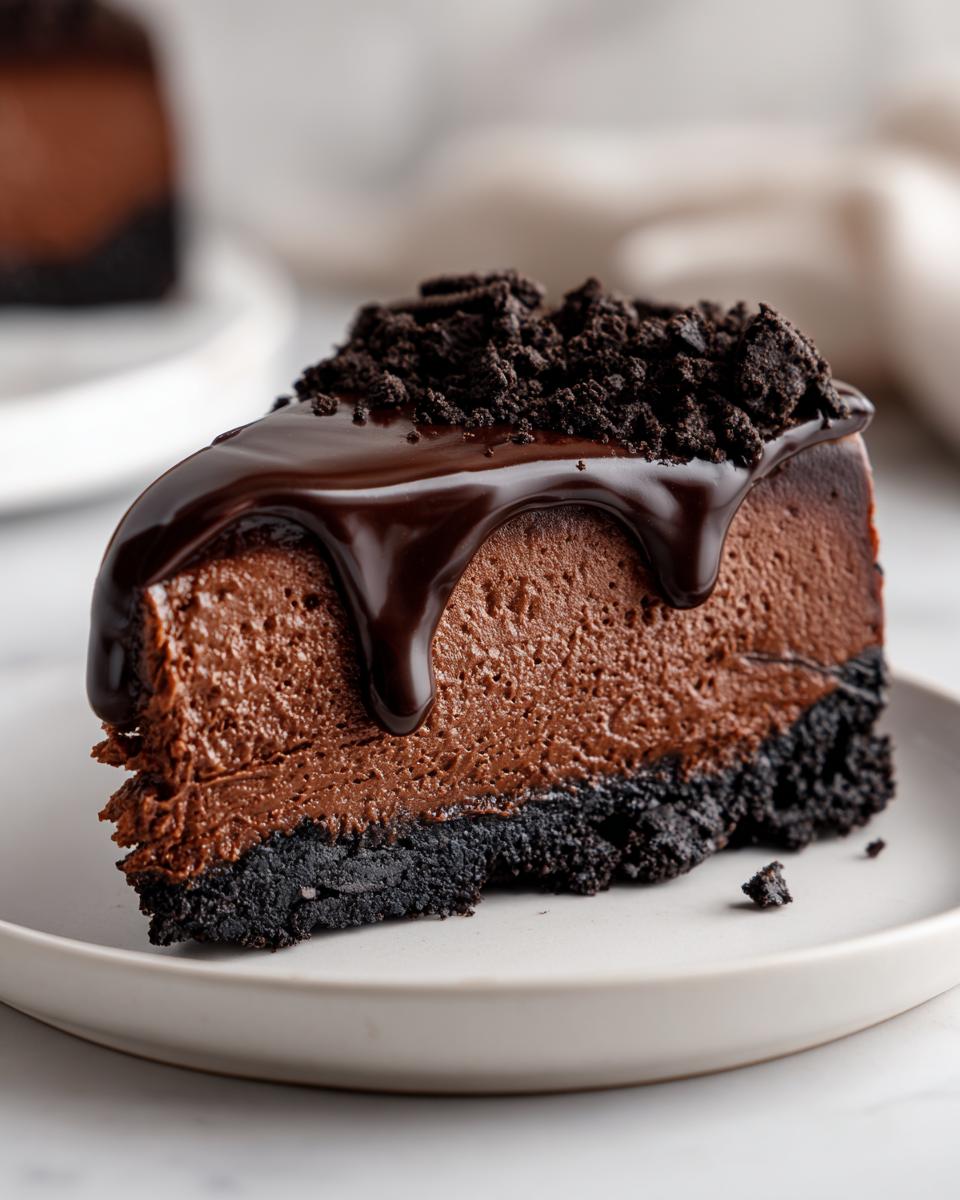 A decadent slice of chocolate cheesecake with a dark cookie crust, topped with chocolate ganache and crushed cookies.