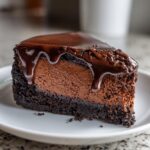 A rich slice of chocolate cheesecake with a thick, dark chocolate crust and a glossy chocolate ganache dripping down the sides.