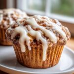 A close-up of a freshly baked cinnamon roll muffin topped with thick white icing dripping down the sides.