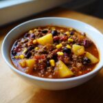 Close-up of a white bowl filled with rich, red cowboy soup featuring ground beef, large potato chunks, black beans, kidney beans, and corn.