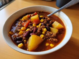 A close-up of a white bowl filled with rich, red cowboy soup featuring ground beef, kidney beans, corn, and chunks of potato.