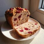 A loaf of homemade cranberry walnut bread, partially sliced, showcasing dried cranberries and walnuts inside.