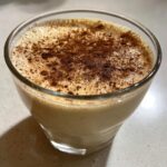 Close-up of a glass filled with creamy coquito recipe, topped with a dusting of ground cinnamon.