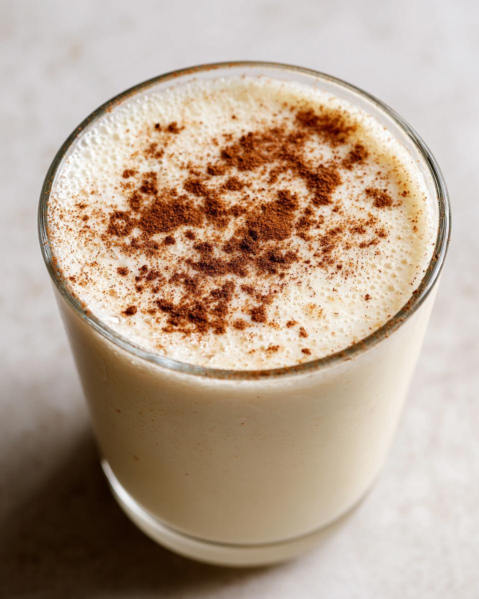 Close-up of a glass filled with creamy coquito recipe, garnished generously with ground cinnamon on top.