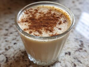 Close-up of a glass filled with creamy coquito recipe, dusted generously with ground cinnamon on top.