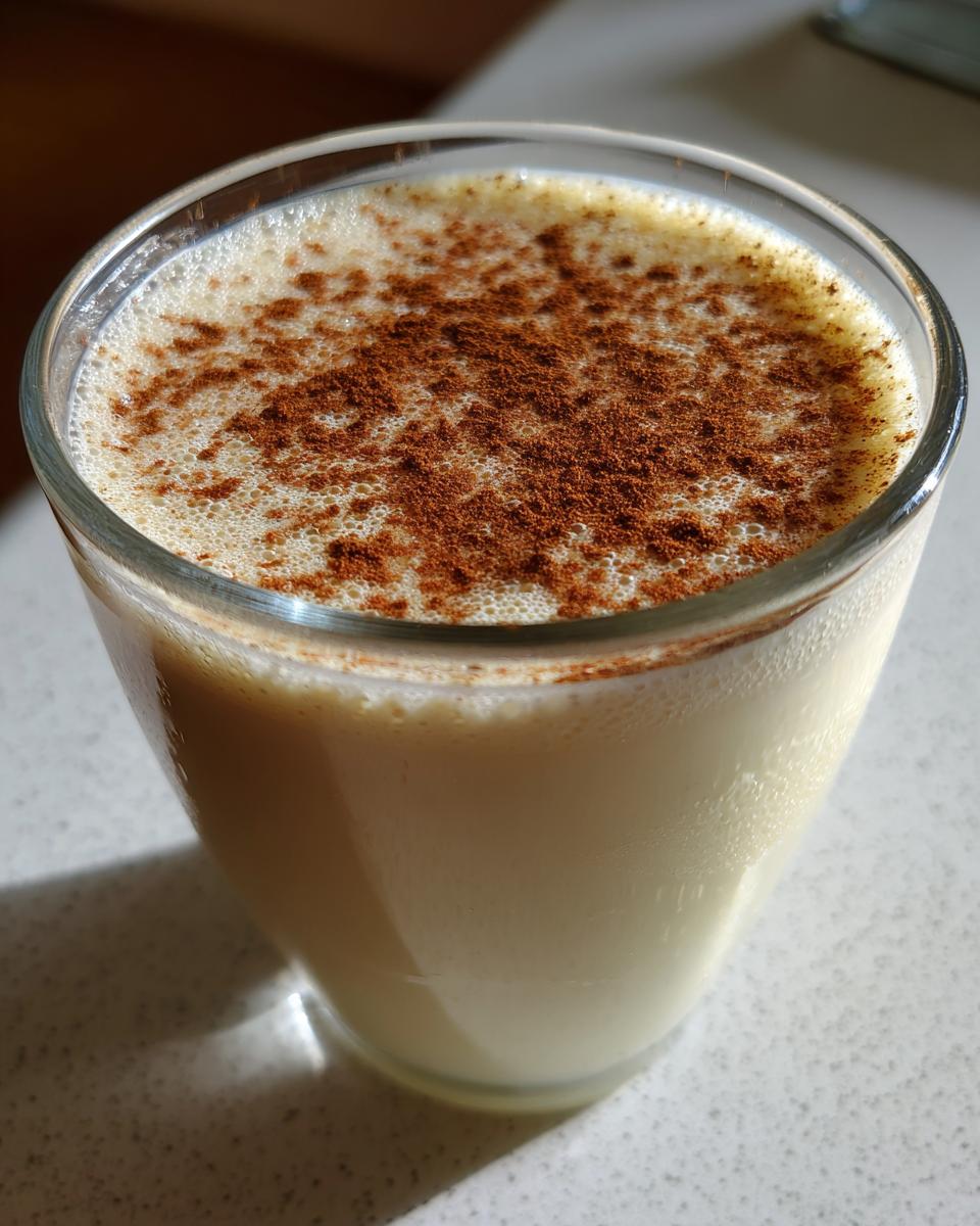 Close-up of a glass filled with creamy coquito recipe, garnished with a dusting of ground cinnamon on top.