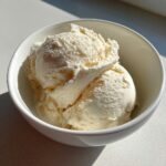 Two scoops of creamy, pale yellow cottage cheese ice cream served in a small white bowl, highlighted by bright sunlight.