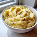A white bowl filled with fluffy garlic mashed potatoes, topped with melted butter and crispy browned garlic bits.
