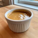 A small white ramekin filled with creamy, rich peanut sauce sitting on a wooden countertop.