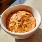 Close-up of a small white bowl filled with thick, creamy orange remoulade sauce, topped with chopped green herbs.