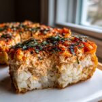 A close-up, cross-section view of a creamy sushi bake slice showing the rice base and savory topping.