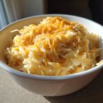 Close-up of a white bowl filled with shredded crockpot cheesy potatoes topped with melted cheddar cheese.
