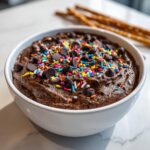 Close-up of rich brownie batter dip topped with colorful sprinkles and chocolate chips, ready to eat.