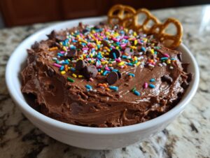 A white bowl filled with rich, chocolate brownie batter dip, topped generously with colorful sprinkles and chocolate chips, garnished with pretzels.
