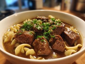 Close-up of a white bowl filled with rich beef noodle soup, featuring large chunks of beef and egg noodles, topped with fresh parsley.