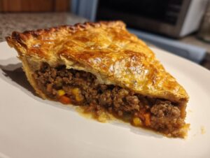 A close-up slice of a homemade meat pie showing a golden, flaky crust and a rich ground beef filling with visible carrots and corn.