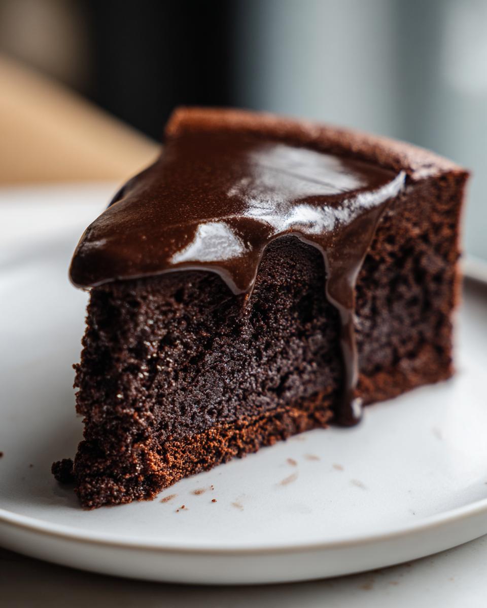 A close-up of a dense slice of flourless chocolate cake topped with glossy, dripping chocolate ganache.