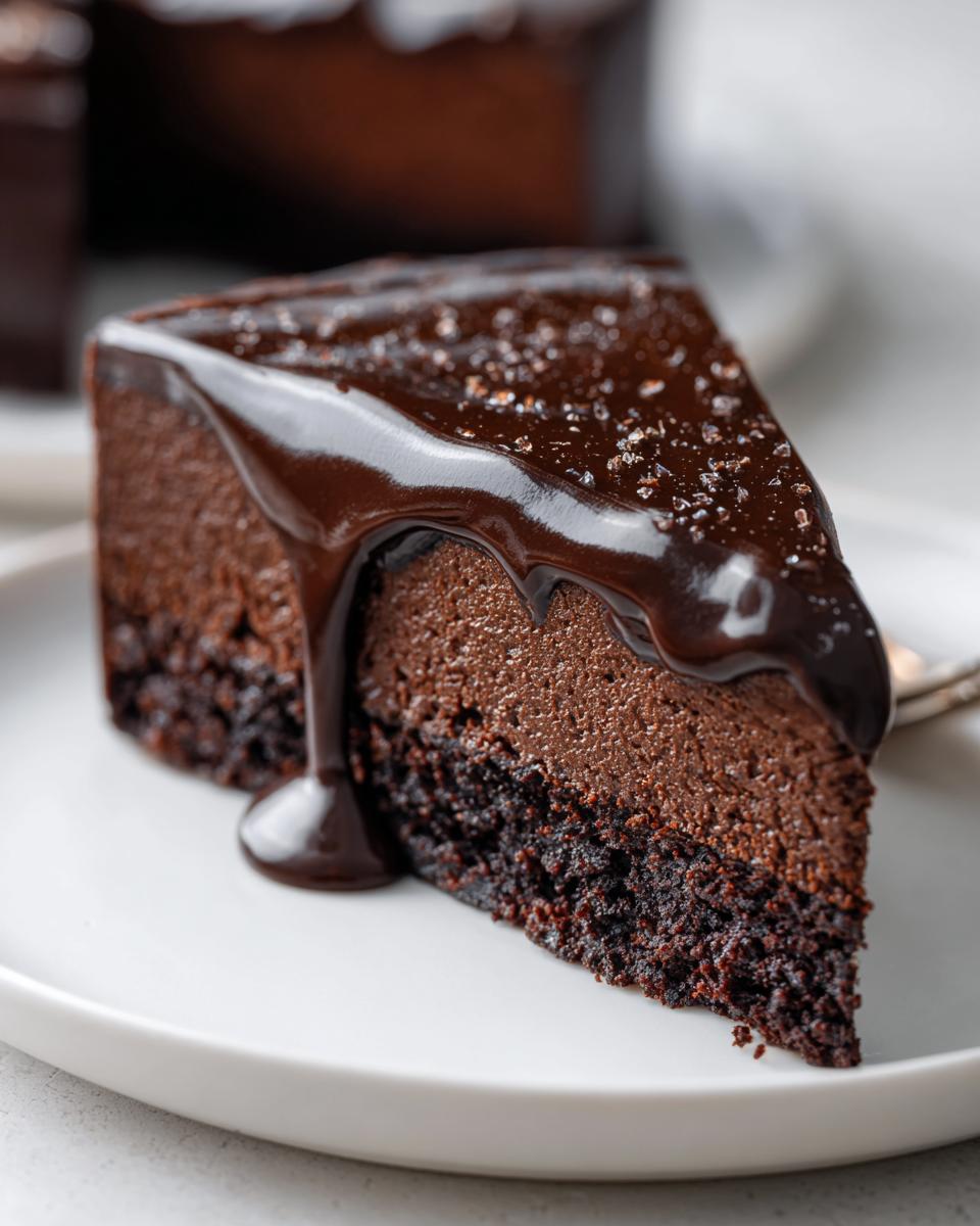 A rich slice of flourless chocolate cake featuring a dense base and a lighter mousse layer, topped with dripping chocolate ganache and sea salt.