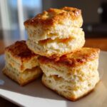 Three tall, flaky buttermilk biscuits stacked on a white plate, showing a golden-brown top.