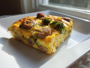 A square slice of fluffy egg bake with sausage and broccoli florets served on a white plate.