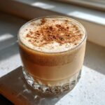 Close-up of a glass filled with creamy coquito, topped with a generous dusting of cinnamon powder.