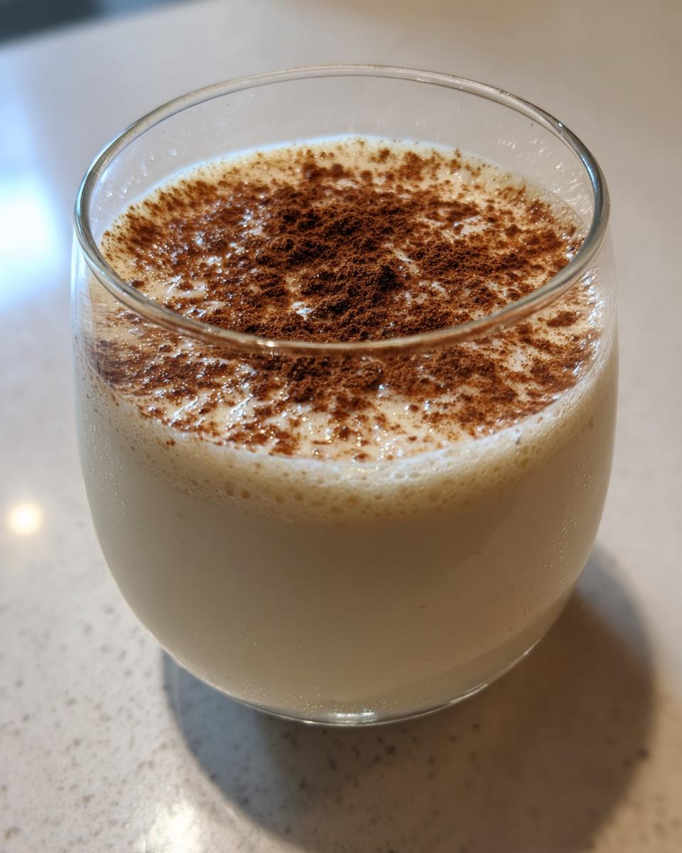 A close-up of a glass filled with creamy coquito, garnished generously with ground cinnamon on top.