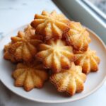 A close-up stack of golden, star-shaped butter cookies sprinkled with sugar on a white plate.