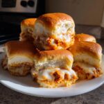 A stack of gooey, cheesy buffalo chicken sliders on a white plate, featuring melted white cheese and orange buffalo sauce.