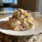 A scoop of creamy greek yogurt chicken salad mixed with diced red onion and green celery on a white plate.