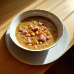 A close-up bowl of hearty ham and white bean soup with carrots and celery, served on a wooden table.