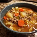 Close-up of a bowl of rich hamburger soup filled with ground beef, carrots, potatoes, and egg noodles.